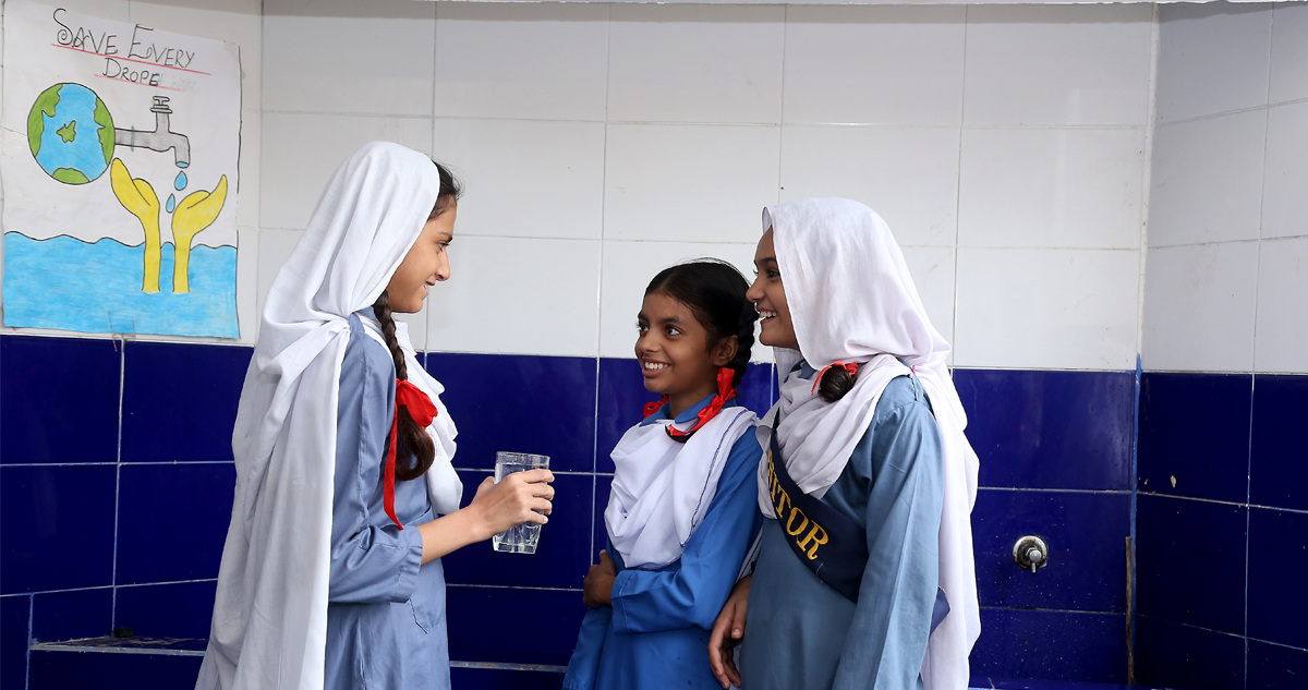 Three young female children next to a sink. One girl holds a glass of water.